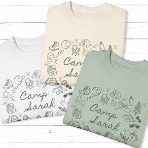 May include: Three t-shirts with a camping theme. The shirts are white, beige, and green. Each shirt has a circle of camping-related illustrations and the text "Camp Sarah".