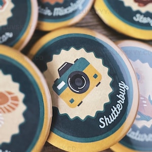 May include: A collection of circular pin-back buttons featuring various graphic illustrations. One prominent button displays a yellow and teal camera icon centered on a tan scalloped background with the text Shutterbug in white script.