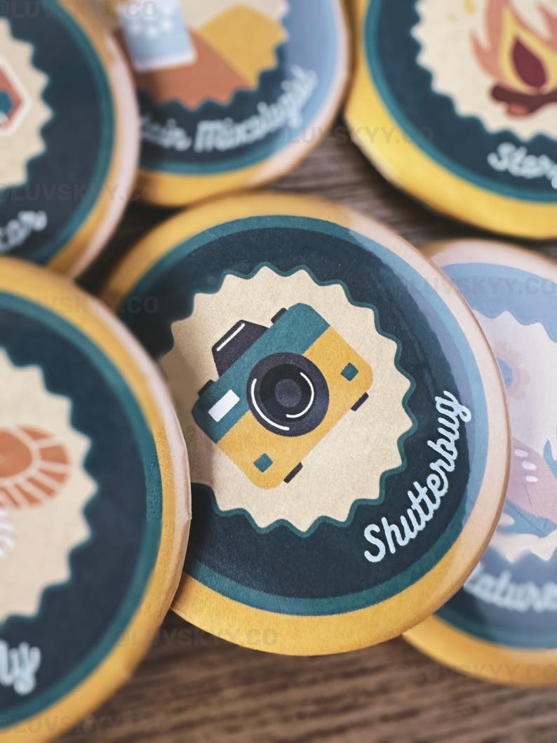 May include: A collection of circular pin-back buttons featuring various graphic illustrations. One prominent button displays a yellow and teal camera icon centered on a tan scalloped background with the text Shutterbug in white script.