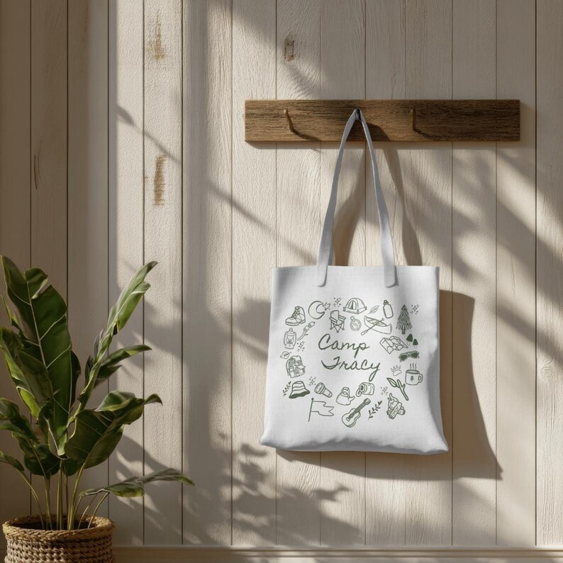 May include: White canvas tote bag with a black and white illustration of camping-related items and the text "Camp Tracy". The bag is hanging on a wooden hook against a white wood panel background.