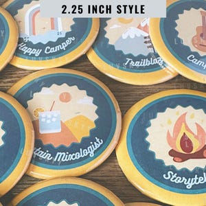 May include: A collection of round pin-back buttons, each 2.25 inch style, featuring various camping-themed illustrations and text including Happy Camper, Trailblazer, Campfire Crooner, Mountain Mixologist, and Storyteller in blue and yellow.