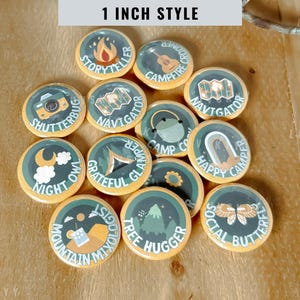 May include: A collection of twelve circular 1-inch buttons featuring camping-themed illustrations and text. Each button displays unique graphics including a campfire, camera, guitar, map, tent, moon, mountain, and butterfly with labels like Storyteller, Navigator, and Tree Hugger.