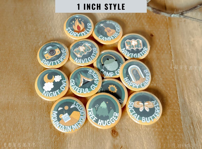 May include: A collection of twelve circular 1-inch buttons featuring camping-themed illustrations and text. Each button displays unique graphics including a campfire, camera, guitar, map, tent, moon, mountain, and butterfly with labels like Storyteller, Navigator, and Tree Hugger.