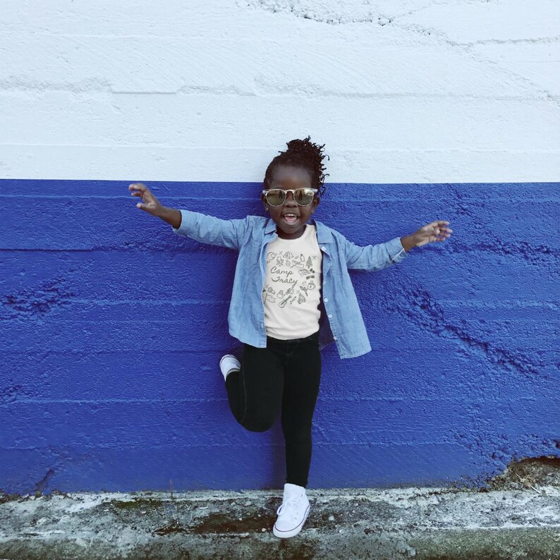 May include: A young person wearing a light blue denim shirt over a white t-shirt with the text "Camp Ready" and black pants. The person is wearing sunglasses and has their arm raised. The background is a blue and white wall.