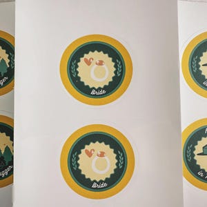 May include: A sheet of six circular stickers with yellow borders and dark green centers. Two stickers feature a mountain graphic with the text Tree Hugger, two show a ring with the text Bride, and two display a hat and badge with Ranger in Training.