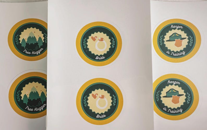 May include: A sheet of six circular stickers with yellow borders and dark green centers. Two stickers feature a mountain graphic with the text Tree Hugger, two show a ring with the text Bride, and two display a hat and badge with Ranger in Training.