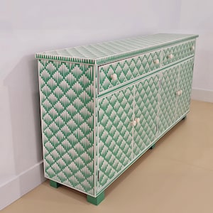 May include: A rectangular sideboard featuring a green and white geometric fan pattern across its entire surface. The furniture piece includes two drawers and four cabinet doors, each fitted with small, round, white knobs and supported by block feet.