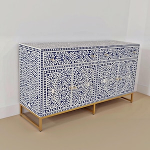 Peut inclure: Un buffet rectangulaire présentant un motif floral incrusté bleu foncé et blanc sur toute sa surface. Le meuble comprend deux tiroirs supérieurs et deux portes de placard inférieures à motifs circulaires, reposant sur un cadre en métal doré.