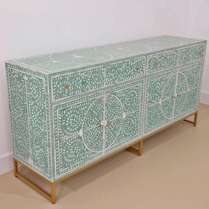 May include: A rectangular sideboard featuring a light green surface with intricate white floral and leaf inlay patterns. The piece includes three top drawers and two lower cabinet doors with circular designs, all supported by a gold metal frame base.