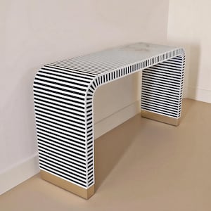 Könnte beinhalten: Ein rechteckiger Konsolentisch mit durchgehendem Wasserfall-Design und einem horizontalen schwarz-weißen Streifenmuster. Der Sockel an beiden Seiten ist mit einem massiven goldfarbenen Metallakzent versehen, der einen Kontrast zur Oberfläche bildet.
