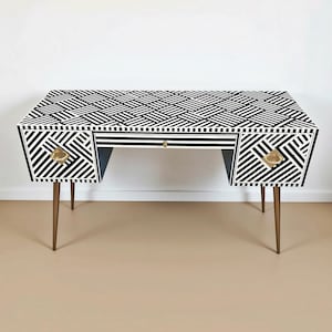 May include: A rectangular desk featuring a black and white geometric striped pattern across the top and front panels. The desk includes a central drawer and two side cabinets with gold-toned hardware, supported by four tapered gold-toned legs.