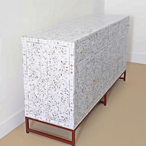 May include: A rectangular sideboard featuring an intricate white and grey floral inlay pattern across its surface. The cabinet includes multiple drawers and doors with small gold-toned metal handles, supported by a simple dark brown metal frame base.