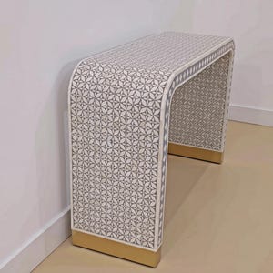 May include: A waterfall console table featuring a repeating geometric star pattern in shades of grey and off-white. The rectangular piece has a smooth, continuous surface and is supported by two solid gold-toned metal bases at the bottom.