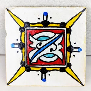 Puede incluir: Azulejo cerámico cuadrado con diseño pintado a mano. El motivo central es un cuadrado rojo con un diseño blanco y azul. El azulejo tiene detalles en amarillo, negro y azul. El azulejo es blanquecino con un aspecto ligeramente envejecido.
