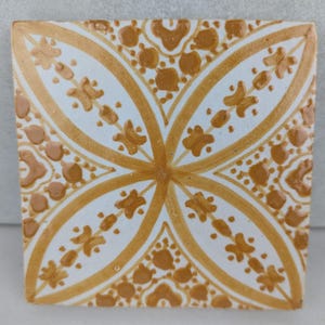 Könnte beinhalten: Quadratische Keramikfliese mit weißem Hintergrund und einem symmetrischen Design in Gold- und Brauntönen. Das Muster weist florale und geometrische Elemente auf und erzeugt ein dekoratives und künstlerisches Aussehen. Die Fliese eignet sich für Heimdekorations- und Designprojekte.