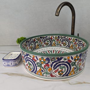 May include: A round ceramic sink with a colorful floral pattern in blue, red, yellow, and green. The sink has a green border and is accompanied by a matching soap dish. A bronze faucet is visible.