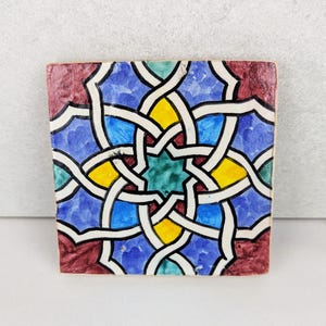 Puede incluir: Azulejo cerámico cuadrado con un patrón geométrico. El diseño presenta líneas blancas entrelazadas sobre un fondo azul, amarillo, verde y granate. El azulejo tiene un aspecto rústico y hecho a mano, con pinceladas y variaciones de color visibles.