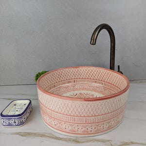 Puede incluir: Un lavabo de cerámica decorativo con un diseño estampado en tonos rosa y blanco. A la izquierda hay un jabonera a juego con diseños azules y blancos. Un grifo de bronce está montado encima del lavabo.