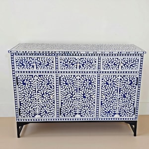 May include: A rectangular sideboard cabinet featuring a deep blue and white floral vine inlay pattern across the top, three upper drawers, and three lower cabinet doors. The piece is supported by four black legs and includes small round knobs.