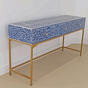 May include: A rectangular console table featuring a blue and white floral inlay pattern across the top and three front drawers. The unit is supported by a thin, gold-toned metal frame with four legs and a horizontal support bar near the base.