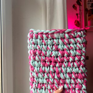 Könnte beinhalten: Eine rechteckige Häkeltasche aus dicken, abwechselnden Strängen aus leuchtend rosa und hellminzgrünem Stoffgarn. Die Tasche weist eine sichtbare Webstruktur und ein kleines, rechteckiges weißes Stoffetikett am oberen Rand mit dem Text "IN THE TUESCH" auf.