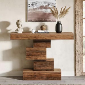 Peut inclure: Une console en bois moderne avec une base géométrique unique. La table est de couleur marron chaud et présente un plateau rectangulaire avec une série de blocs rectangulaires empilés formant la base. Des objets décoratifs sont posés sur la table.