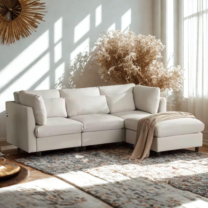 May include: A cream-colored sectional sofa with a matching ottoman. The sofa has several large, fluffy cushions and sits on short, dark legs. A beige throw blanket is draped over the ottoman. The sofa is placed on a patterned rug in a sunlit room.