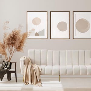 Op de afbeelding: Een moderne woonkamerscène met een crèmekleurige bank met gouden poten, een beige plaid en drie ingelijste abstracte kunstprints in neutrale tinten. Een houten stoel met een donkergroene deken en gedroogde planten in een vaas maken de inrichting compleet.