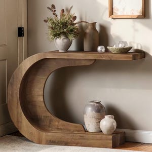 Könnte beinhalten: Eine Holzkonsolentisch mit einem einzigartigen, geschwungenen Design. Der Tisch hat eine warme braune Farbe und eine Ablagefläche oben. Dekorative Vasen und eine Schale mit Kerzen sind auf der Ablage ausgestellt. Der Tisch ist eine stilvolle Ergänzung für jeden Wohnraum.