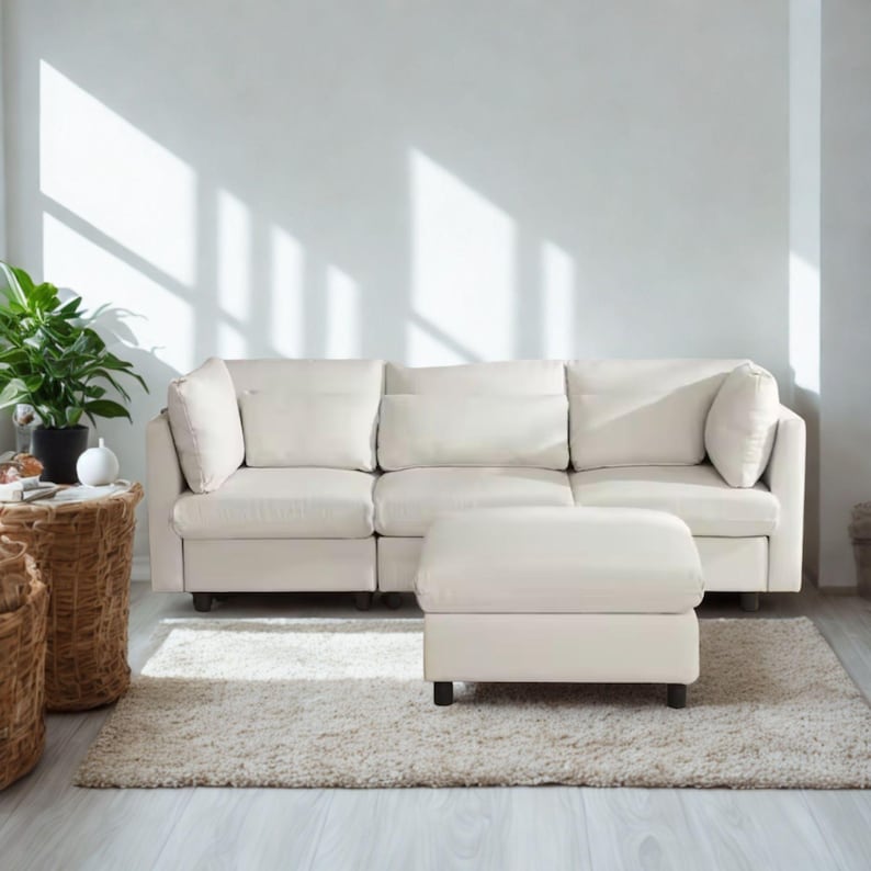 May include: A cream-colored sectional sofa with matching ottoman sits on a shaggy rug. The sofa has three sections with large, square cushions and pillows. A potted plant and woven side tables are on the left.