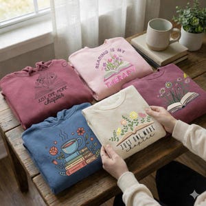 May include: Several sweatshirts and a t-shirt with book-themed designs. The sweatshirts are in shades of pink, burgundy, and blue. The t-shirt is cream-coloured. Each garment features floral and book graphics with phrases like "Reading is my therapy" and "Just one more chapter."