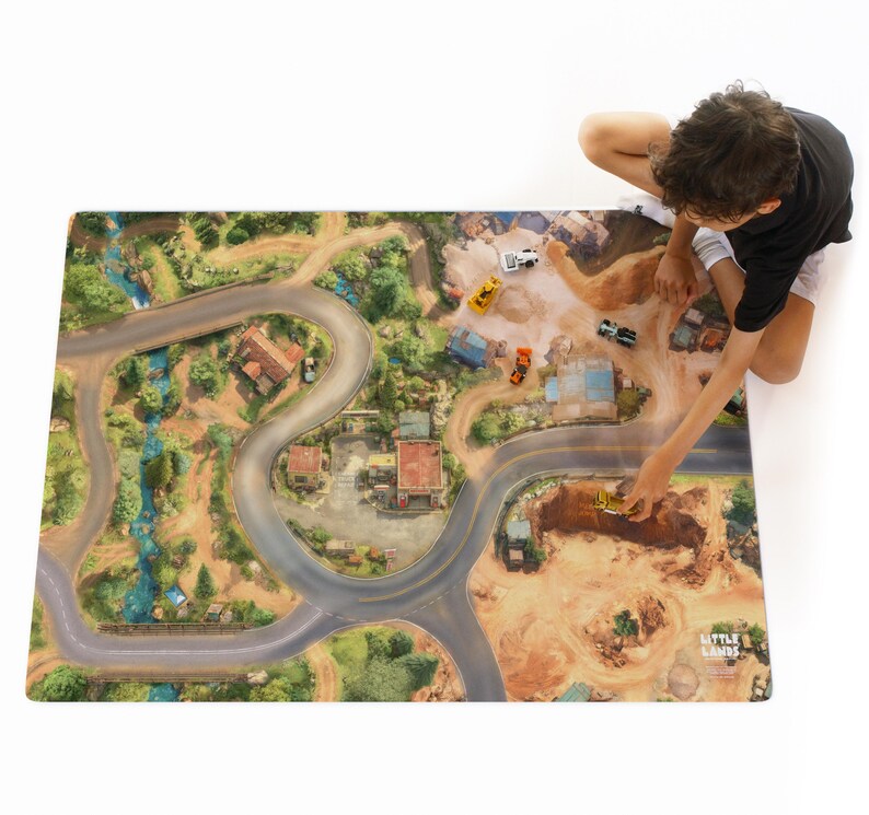 May include: A rectangular play mat featuring a printed aerial view of a landscape with winding roads, a blue stream, green trees, and buildings. The mat includes small toy construction vehicles and the text Little Lands in the bottom corner.