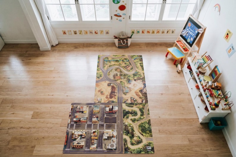 May include: A rectangular play mat featuring a printed design of a city street grid, winding roads, and natural landscapes. The mat is laid out on a light-coloured wooden floor in a room with a white storage unit, a wooden easel, and toys.