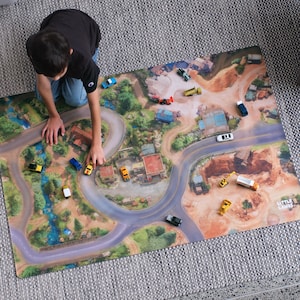 May include: A rectangular play mat featuring a printed aerial view of a landscape with winding roads, buildings, and natural terrain. Several small toy vehicles are scattered across the surface, which includes the text Little Lands in the corner.