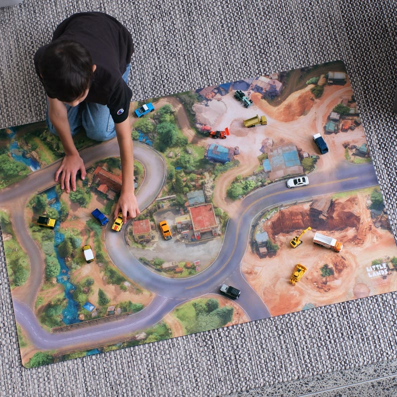 May include: A rectangular play mat featuring a printed aerial view of a landscape with winding roads, buildings, and natural terrain. Several small toy vehicles are scattered across the surface, which includes the text Little Lands in the corner.
