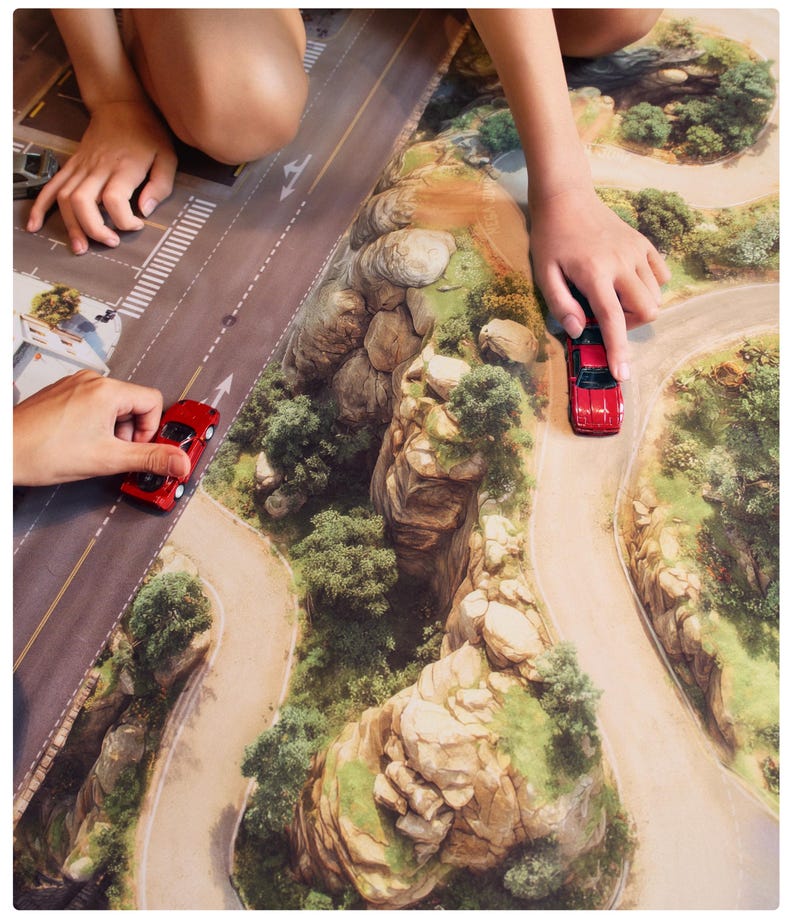 May include: A play mat featuring a printed landscape with a paved road, winding dirt paths, and rocky terrain with green trees. Two small red toy cars are positioned on the mat, one on the paved road and one on the dirt path.