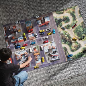 May include: A rectangular play mat featuring a printed city street grid on the left and a winding mountain road on the right. Numerous small toy cars in various colours are positioned across the urban and natural landscape illustrations.