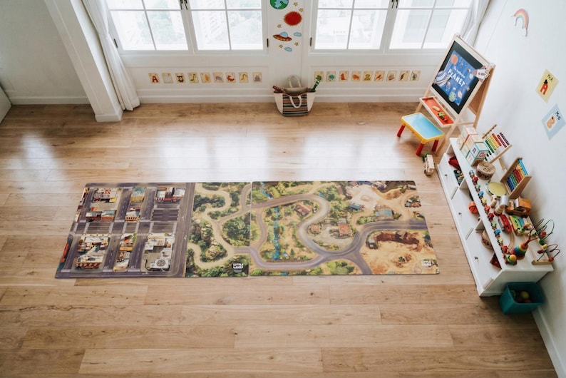 May include: A rectangular play mat featuring a printed design of a city grid on the left and a winding road through a natural landscape on the right. The mat is laid flat on a wooden floor, showcasing detailed illustrations of streets and terrain.