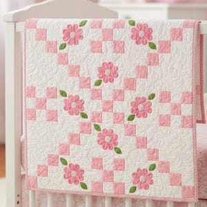 Op de afbeelding: Een wit en roze babyquilt met een bloemenontwerp. De quilt heeft een dambordpatroon met roze vierkanten en geborduurde roze bloemen met groene bladeren. De quilt is over een wit babybedje gedrapeerd.