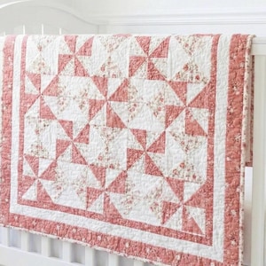 May include: A quilt featuring a pinwheel pattern composed of solid dusty rose and white floral fabric triangles. The quilt has a wide border in a matching dusty rose floral print and is finished with white fabric borders and intricate quilting.