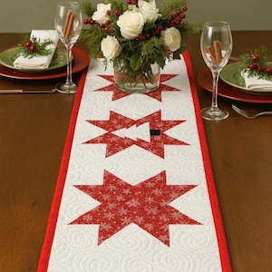 May include: A quilted table runner with a white background and red star pattern. The runner has a red border and is decorated with a floral centerpiece, wine glasses, and plates.