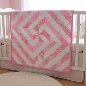 May include: A handmade baby quilt in a crib. The quilt features a geometric design with alternating white and pink fabric strips. The pink fabric includes solid, dotted, and floral patterns. The quilt is trimmed with a pink border.