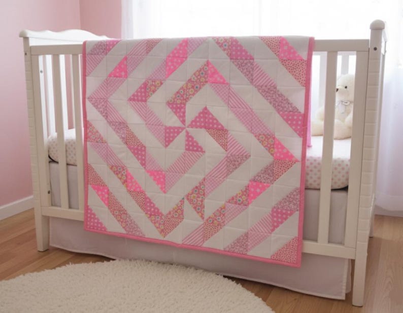 May include: A handmade baby quilt in a crib. The quilt features a geometric design with alternating white and pink fabric strips. The pink fabric includes solid, dotted, and floral patterns. The quilt is trimmed with a pink border.