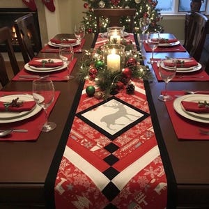 May include: A Christmas table setting with a red and white patterned table runner featuring a reindeer design. The table is set with red placemats, white plates, and wine glasses. A lit Christmas tree and candles create a festive atmosphere.
