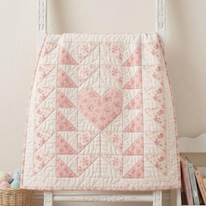 May include: A quilted blanket featuring a central heart motif composed of pink floral fabric patches. The design incorporates alternating cream and pink floral triangular segments arranged in a geometric pattern with visible stitching throughout the fabric.