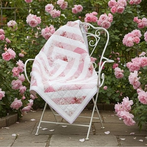 May include: A white metal chair with a pink and white quilt draped over it, set in a garden of pink roses. The quilt features a geometric design with stripes and floral patterns, and is bordered with white fabric.