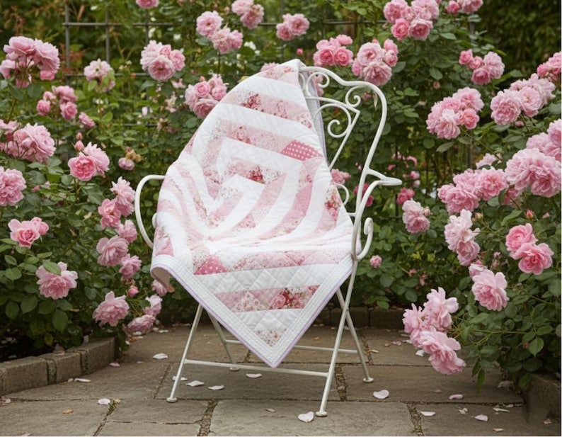 May include: A white metal chair with a pink and white quilt draped over it, set in a garden of pink roses. The quilt features a geometric design with stripes and floral patterns, and is bordered with white fabric.