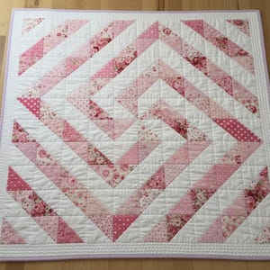 May include: A handmade quilt with a white background and a pink and white chevron pattern. The quilt features various shades of pink fabrics, including floral and polka dot designs. The quilt is bordered with a light purple trim.