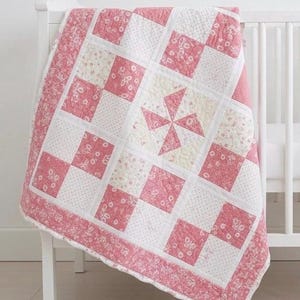 Puede incluir: Una colcha de patchwork con una cuadrícula repetitiva de cuadrados alternos con motivos florales rosas y cuadrados blancos con puntos. El cuadrado central muestra un diseño de molinillo en rosa y crema. Borde ancho con motivos florales.