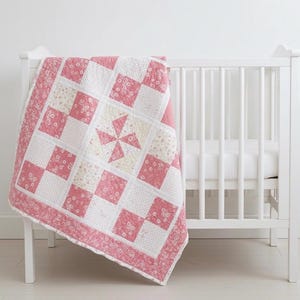 以下が含まれることがあります： ピンクと白のパッチワークキルト。風車模様で、白いベビーベッドに掛けられています。キルトは花柄と無地の正方形で構成され、花柄の縁取りがあります。ベビーベッドには垂直のスラットがあります。